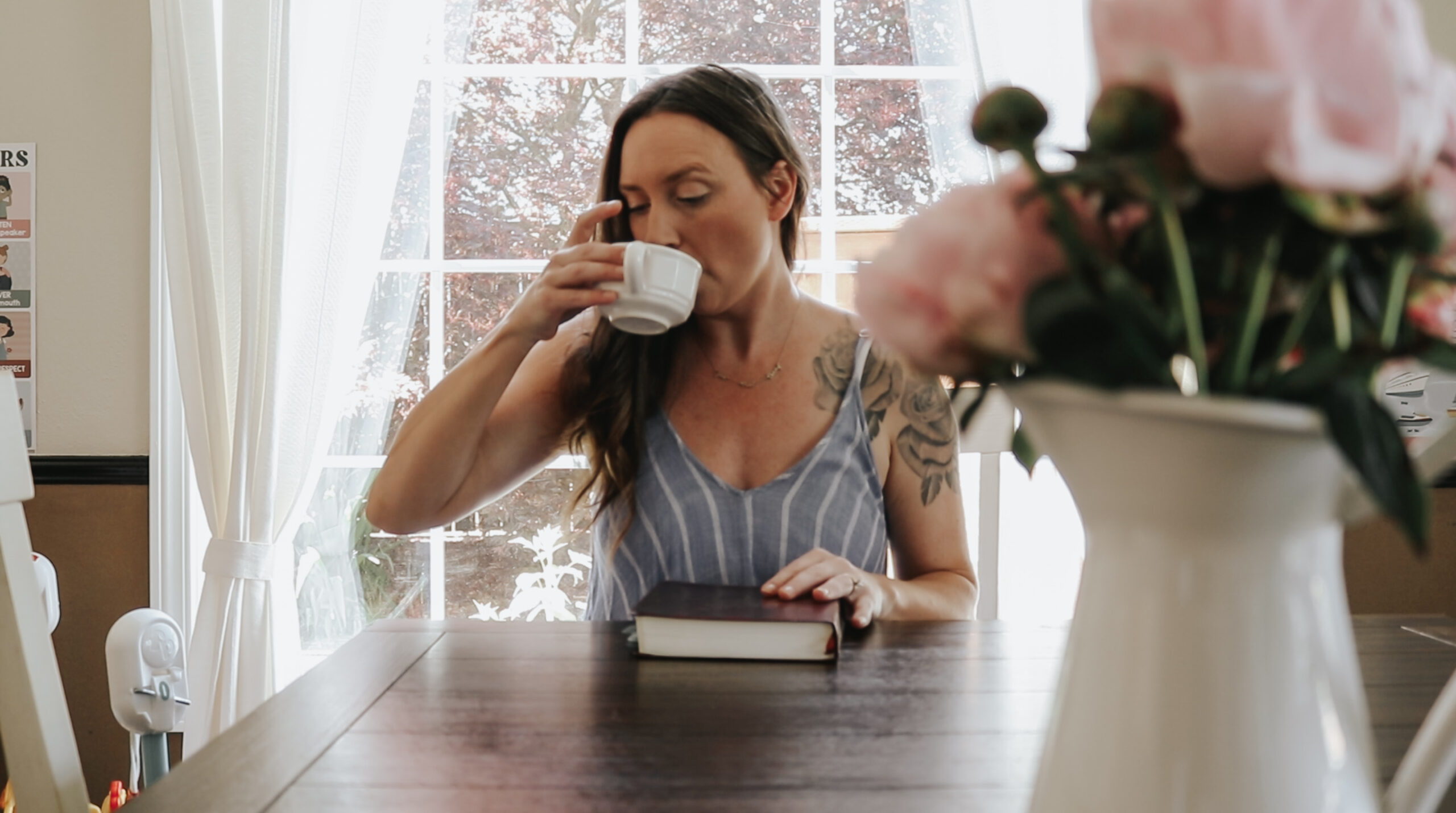 Christian mom and homemaker sits at the table with a white vase full of pink peonies. She is reading her bible and drinking a cup of coffee. It is daytime and sunny.